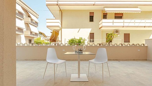 A terrace with a table and two chairs, a tiled floor, and a building with balconies in the background. - Homiday - Albatros 2, Two Bedroom Apartment on the Ground Floor (Alba Adriatica)