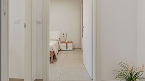 A narrow hallway leading to a bedroom with a bed, bedside table, and a small plant. - Homiday - Albatros 3, Two Bedroom Apartment on the Ground Floor (Alba Adriatica)