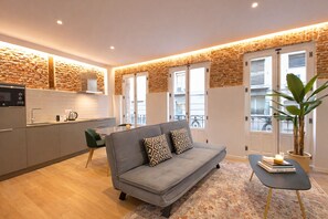 Ein modernes Wohnzimmer mit einem grauen Sofa, einem kleinen Tisch mit Büchern, einer Topfpflanze und einer Küchenzeile mit Mikrowelle und Wasserkocher.