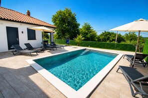 Ein rechteckiger Swimmingpool mit Fliesen umgeben, mit Liegestühlen und Sonnenschirmen, im Hintergrund ein weißes Gebäude und ein grüner Heckenstreifen.