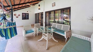 A covered patio with a glass table and white chairs, a hanging hammock, and a view through large windows. - Casa blu 4BHK Spacez Pool & Garden Villa (Bengaluru)