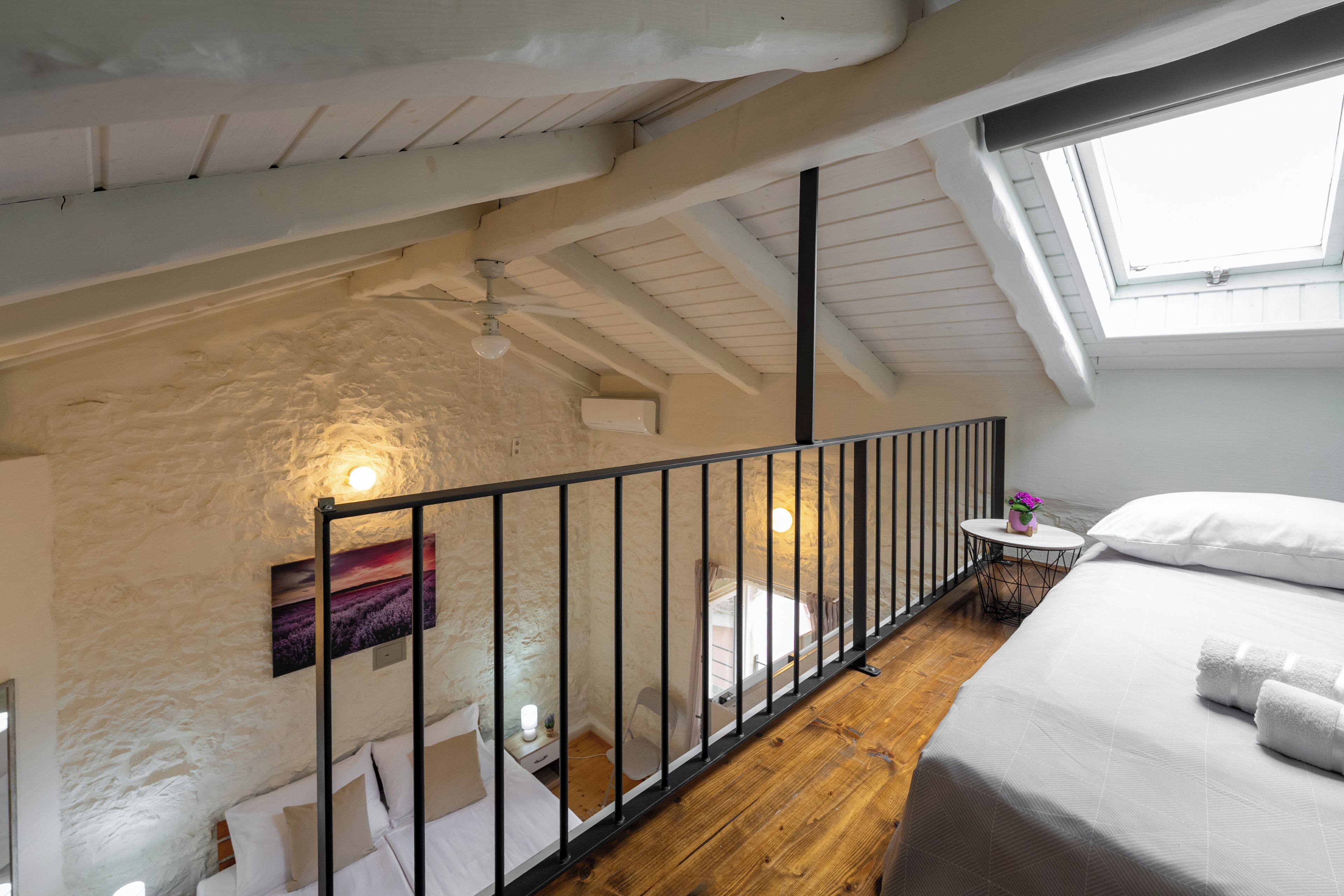 Ein Schlafzimmer mit schrägem Dach, einem Oberlicht, einem Bett mit weißen Leinen, einem kleinen runden Tisch mit einer Vase und einer weißen Wand mit einem Bild.