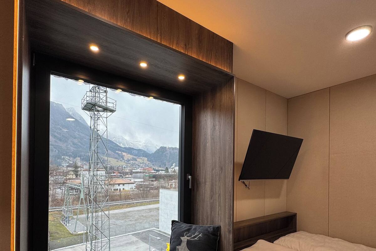 Un dormitorio con un ventanal grande, una cama, televisor montado y vistas a montañas y líneas eléctricas.