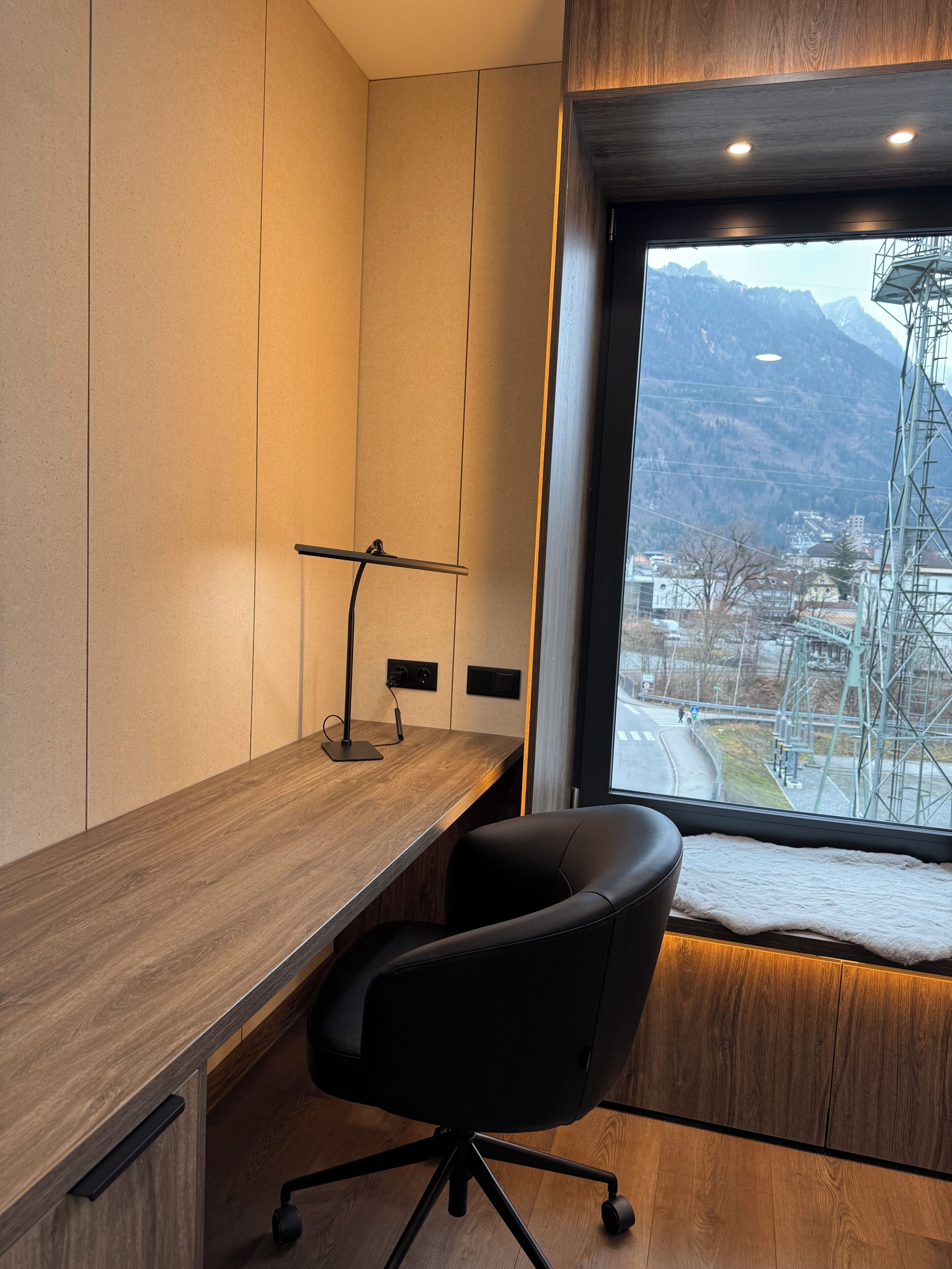 Een moderne kantoorruimte met een houten bureau, een zwarte bureaustoel en uitzicht op bergen door een raam.
