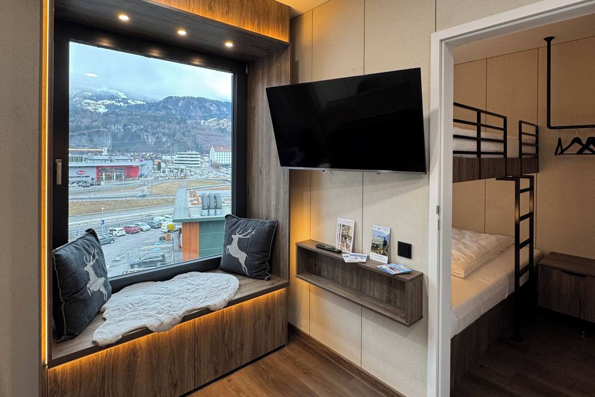 Una habitación moderna con un gran ventanal, un televisor montado, una cama litera y estanterías empotradas.