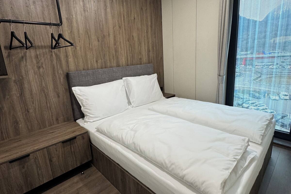 Una habitación de hotel moderna con una cama grande, un cabecero de madera, una mesita de noche pequeña y vistas a un paisaje urbano a través de un ventanal amplio.