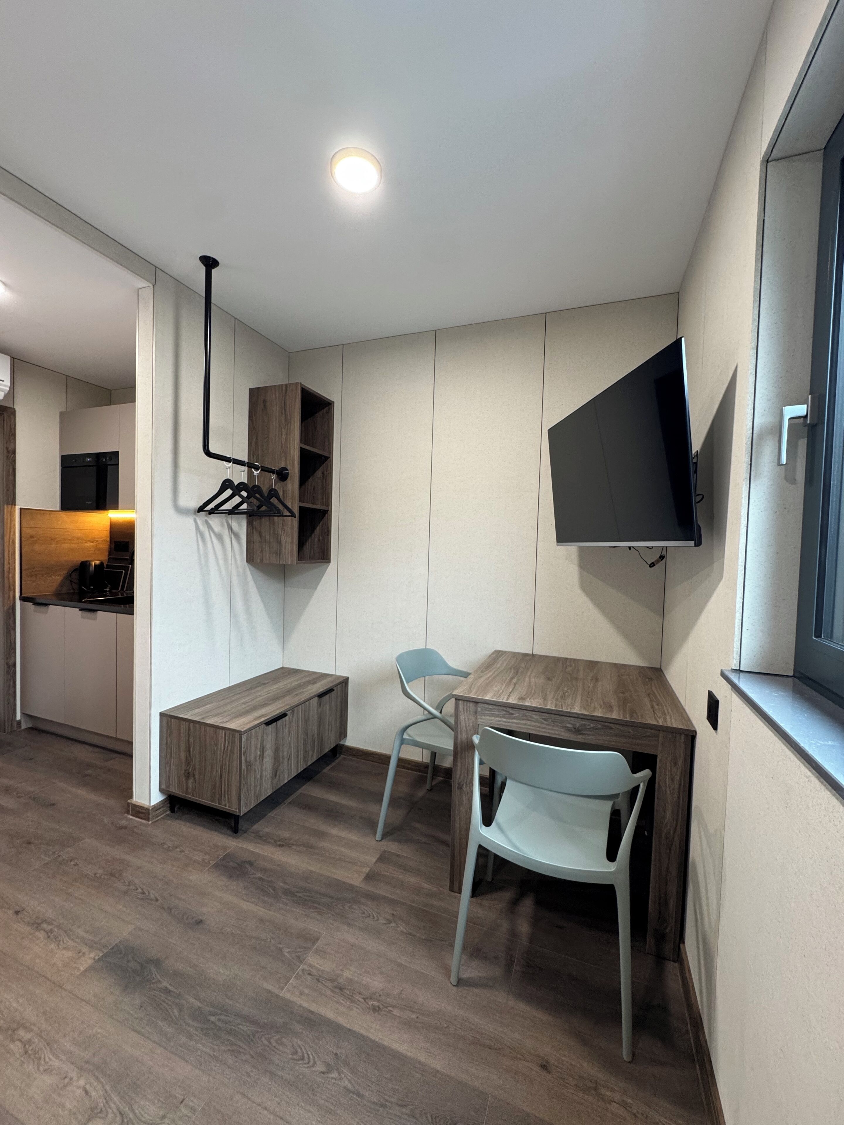 Une cuisine moderne dotée d’un four encastré, un coin repas avec une table en bois et deux chaises, et un téléviseur fixé au mur.