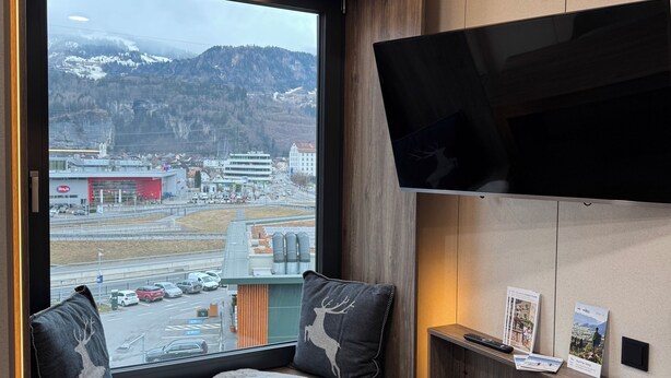 A modern room with a large window, a mounted TV, and a wooden bench with a white fur throw. - Cube Alpine Stay (Buers)