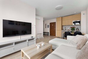 A modern living room with a flat-screen TV, a sofa, a coffee table, and a kitchen area with wooden cabinets and a microwave.