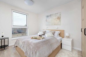 A bedroom with a large window, a bed with a white bedspread, a bedside table with a lamp, and a painting on the wall.