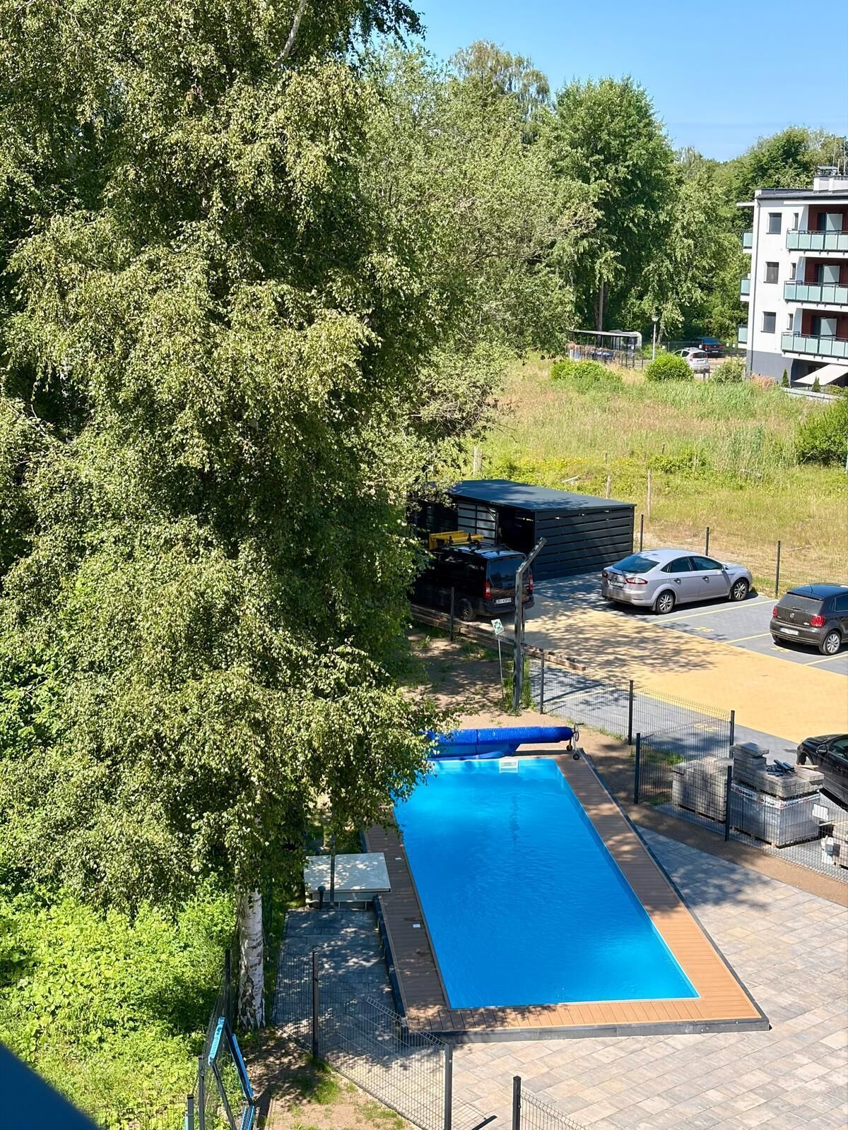 A swimming pool with a wooden deck, surrounded by a fence, and a parking area with cars.