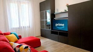 Un salon avec un canapé rouge, un téléviseur affichant le logo de Prime, et une fenêtre avec des rideaux blancs.