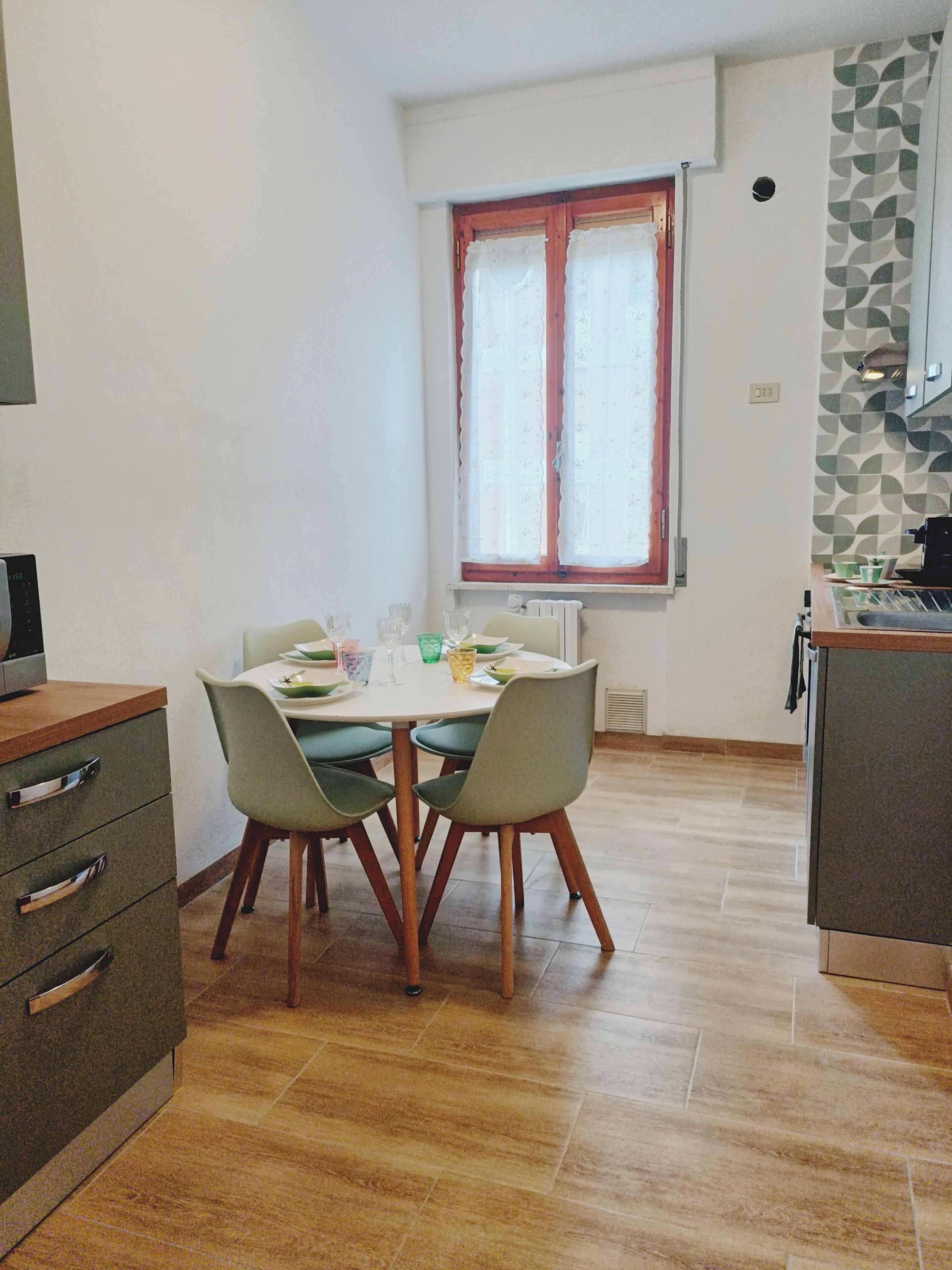 A dining area with a table set for four, a window with curtains, a kitchen area with a refrigerator, and a cabinet with drawers.