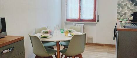 A dining area with a table set for four, a window with curtains, a kitchen area with a refrigerator, and a cabinet with drawers.