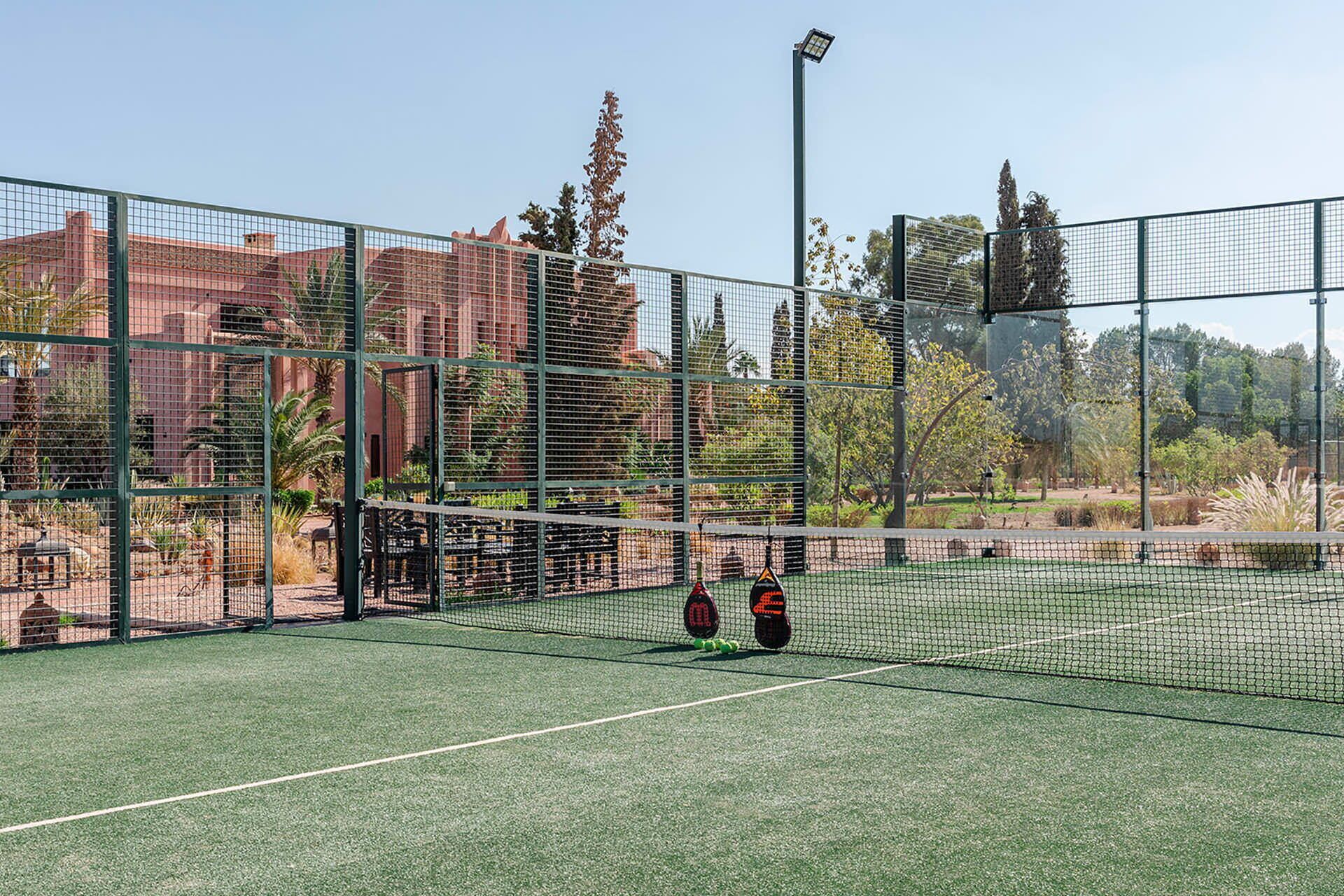 Una cancha de tenis al aire libre con red, rodeada por una valla y árboles al fondo.