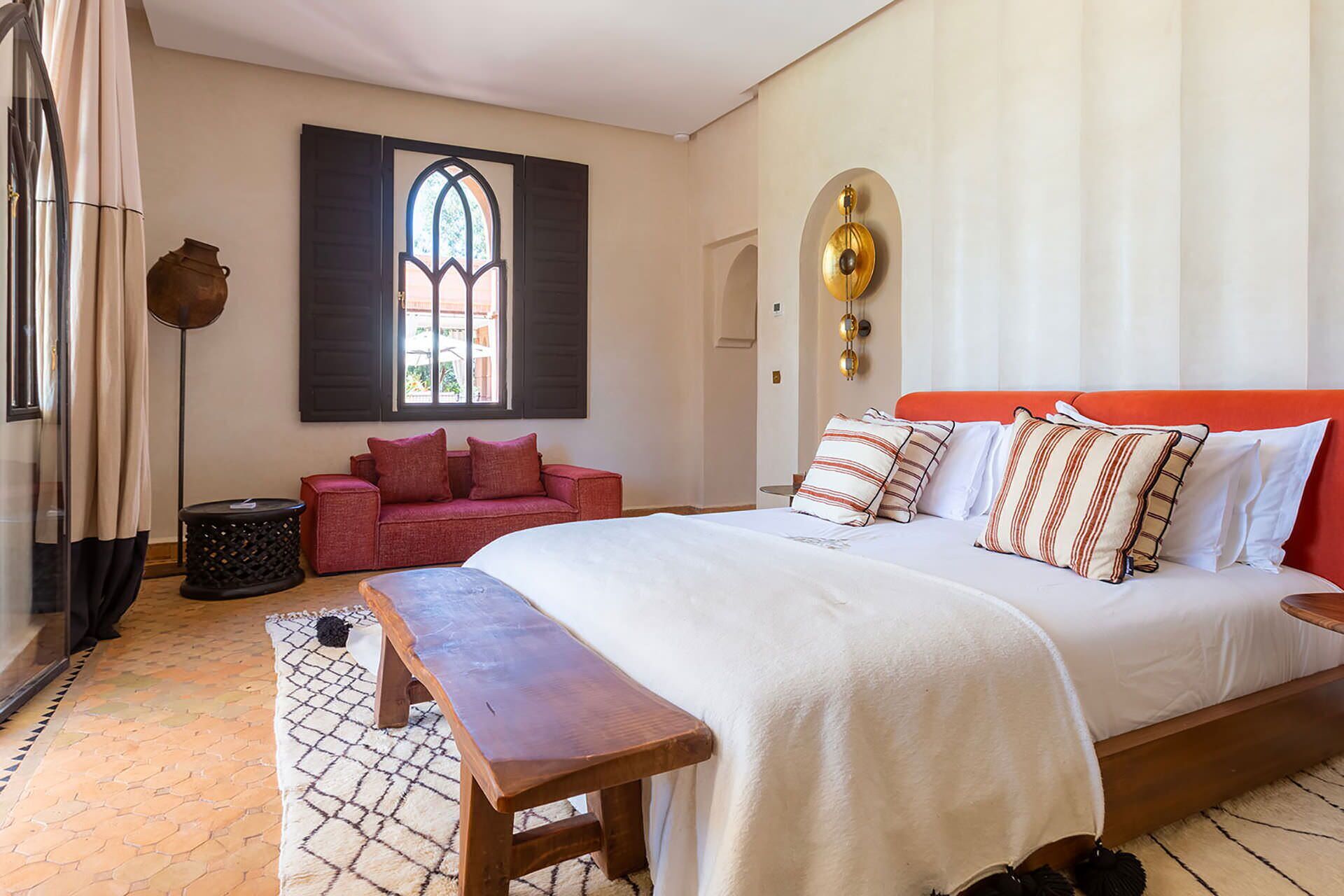 Un dormitorio con una cama grande, un banco, un sofá rojo, una ventana con contraventanas y una lámpara fijada a la pared.