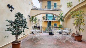 A courtyard with a table and chairs, potted plants, and a building in the background. - Marchese Room with Shared Terrace (Pietra Ligure)