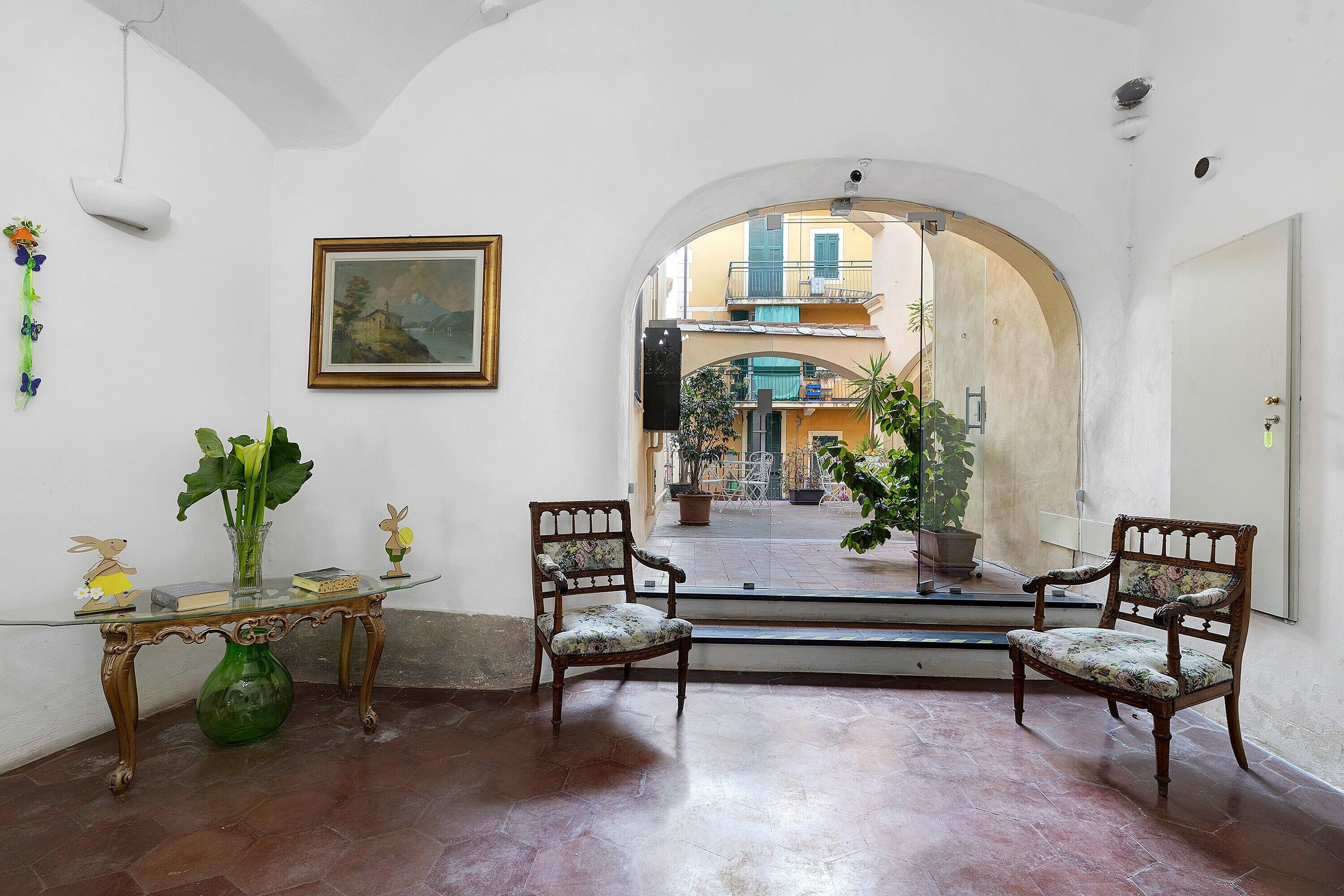 A room with a glass-top table, two chairs, a vase, a framed picture, and an arched doorway leading to an outdoor area with potted plants.