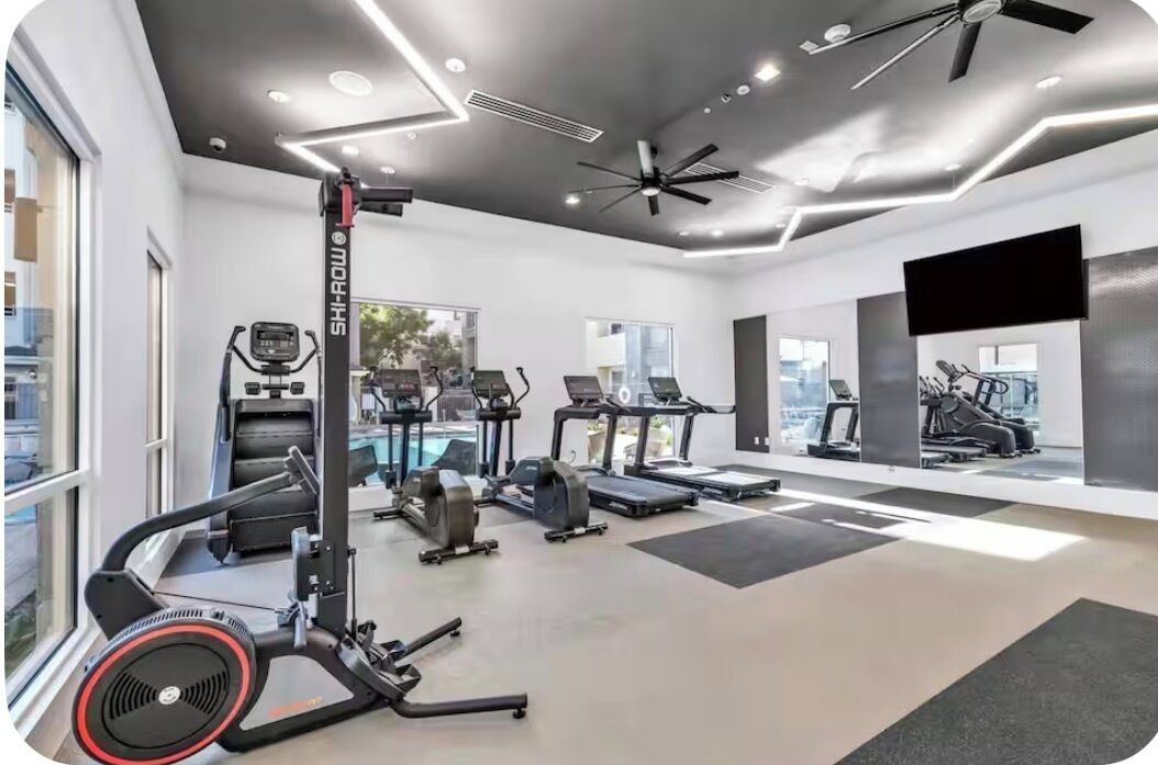 Un gimnasio moderno con varias máquinas de cardio, un ventilador de techo y ventanas amplias.