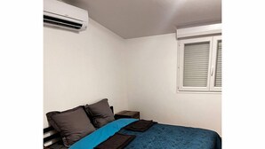 Une chambre à coucher avec un lit, des oreillers, une couverture bleue, un climatiseur, une fenêtre avec des stores et un plancher en bois.
