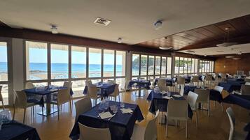 Un restaurant où les tables sont dressées pour le service, de grandes fenêtres donnant sur la plage et un plafond doté d’un éclairage encastré.
