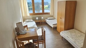 A small, tidy room with a wooden table, two beds, a radiator, and a window with a view of a lake. - Hotel Chytrov (Volfířov)