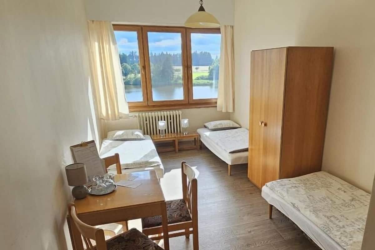 Une petite pièce propre et bien rangée, avec une table en bois, deux lits, un radiateur et une fenêtre donnant sur un lac.