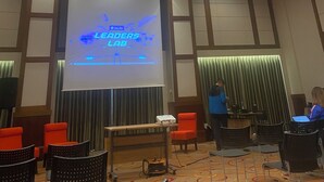 Ein Konferenzraum mit projiziertem Bildschirm, der 'InLife Leaders Lab' anzeigt, einer abgehängten Deckenbeleuchtung und einem Tisch mit Stühlen.