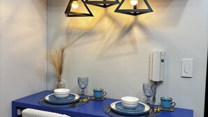Ein Essbereich mit blauem Tisch, drei Pendelleuchten, weißen Stühlen und einer Vase mit Trockenpflanzen.