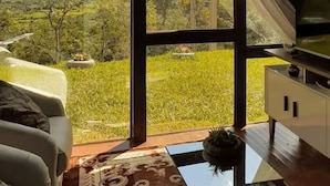 Ett rum med ett stort fönster, en soffa, ett bord och utsikt över ett grönskande landskap med berg.