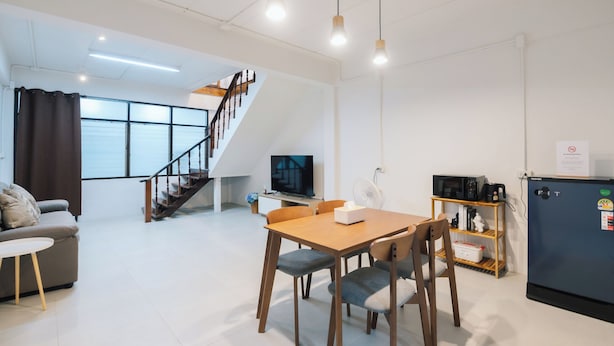 A modern living room with a dining area, a staircase, a flat-screen TV, a refrigerator, and a fan.
