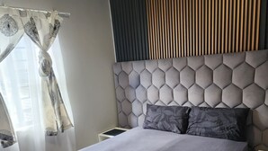 Ein modernes Schlafzimmer mit einem großen Bett, einem Kopfteil mit geometrischem Muster, einem Nachttisch und einem Fenster mit Vorhängen.