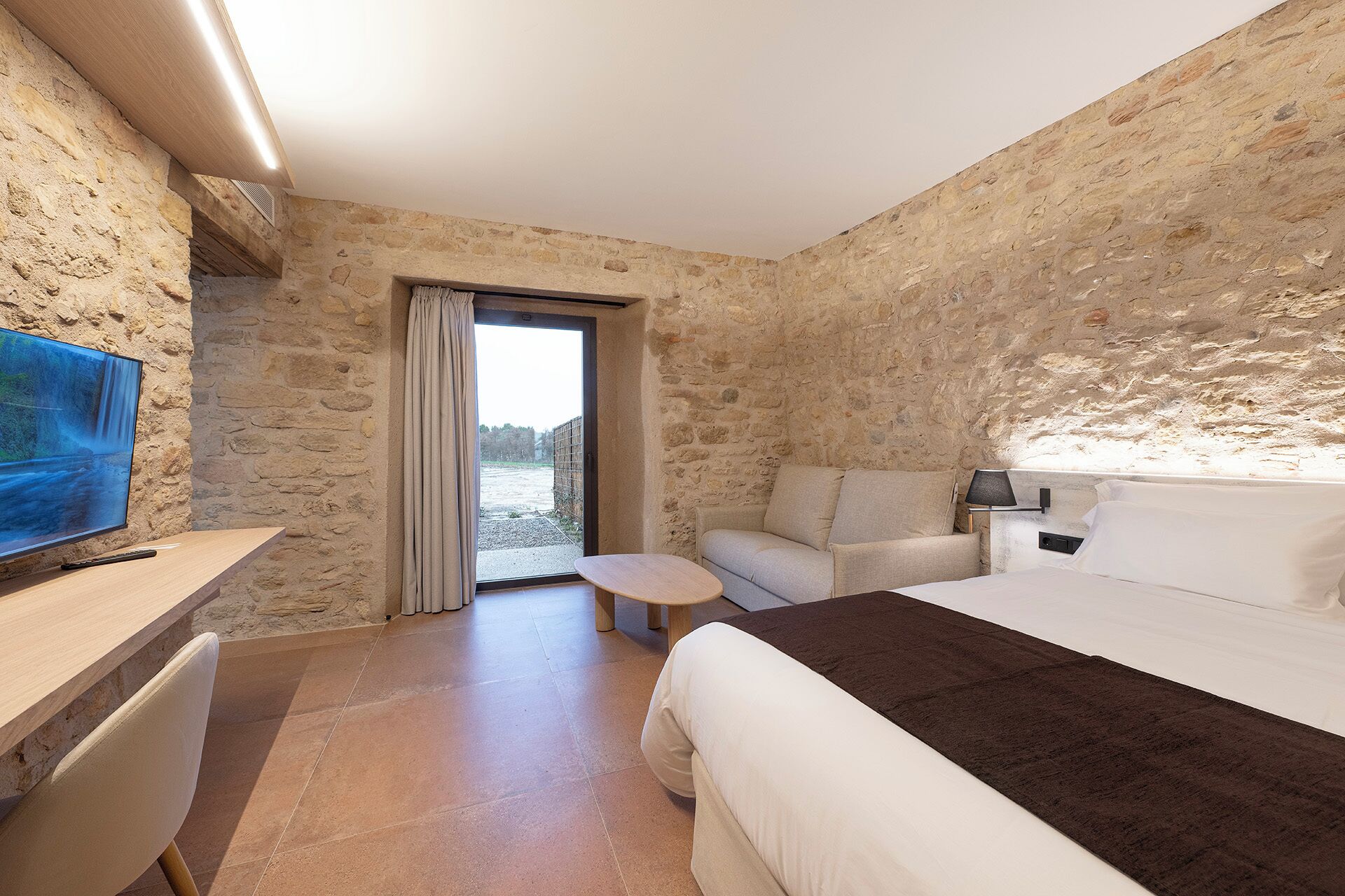 A hotel room with stone walls, a bed, a desk with a TV, a chair, and a small table.