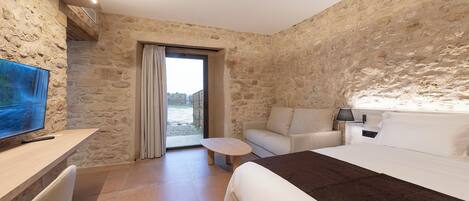 A hotel room with stone walls, a bed, a desk with a TV, a chair, and a small table.