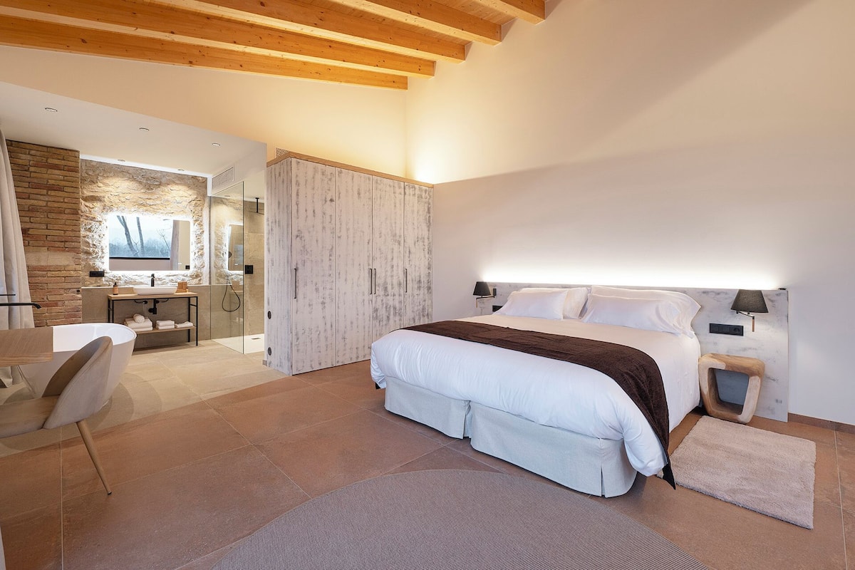 A modern bedroom with a large bed, a wooden ceiling, a stone wall, and a bathroom with a stone shower.