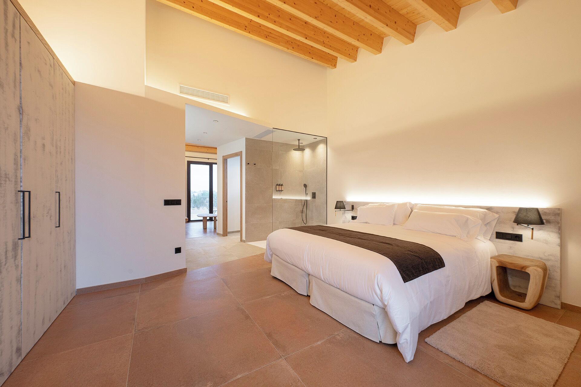 A modern bedroom with a large bed, a bedside table, a mirror, and a shower area.