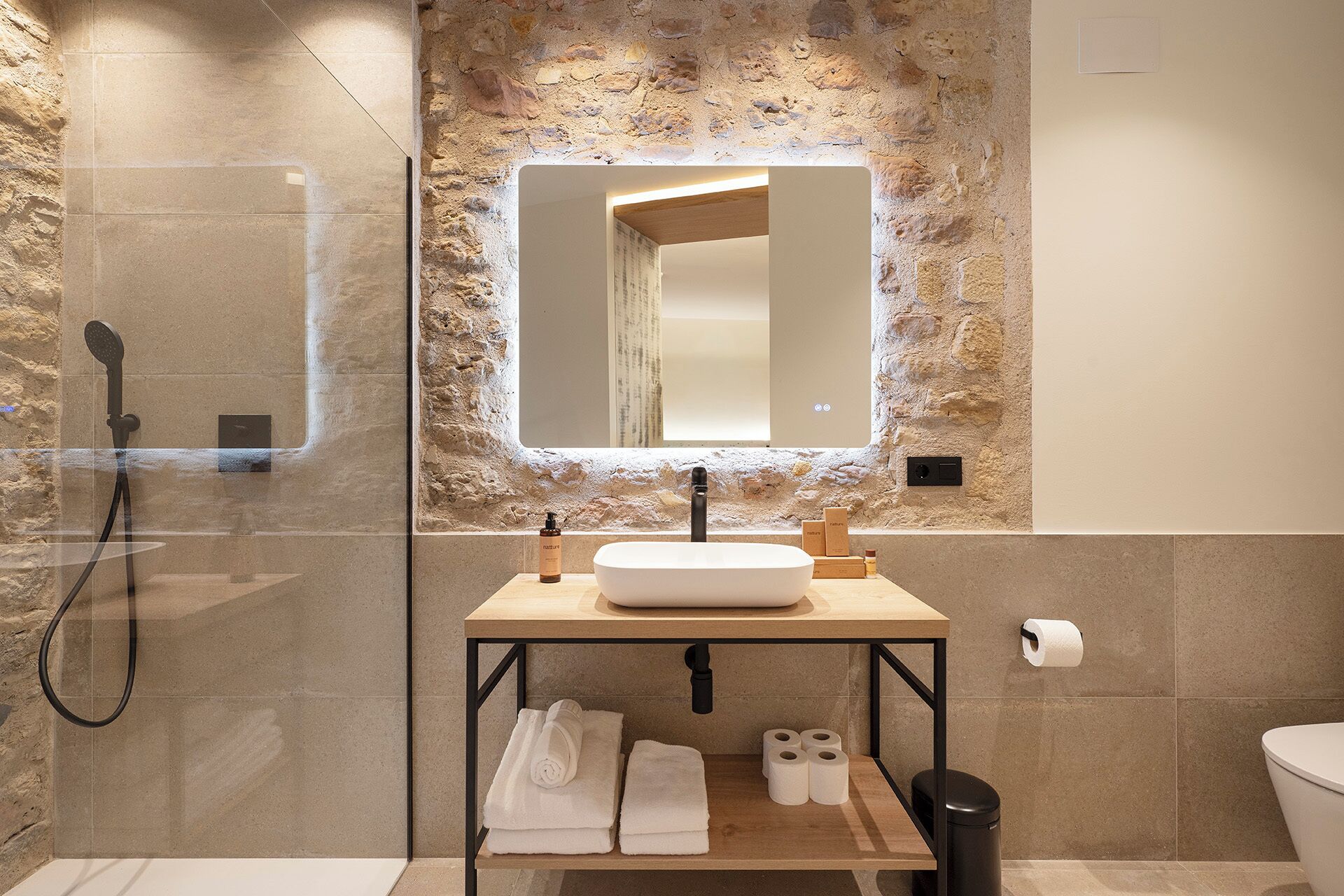 Une salle de bain avec un mur en pierre, une cabine de douche en verre, un meuble-lavabo en bois et un miroir.