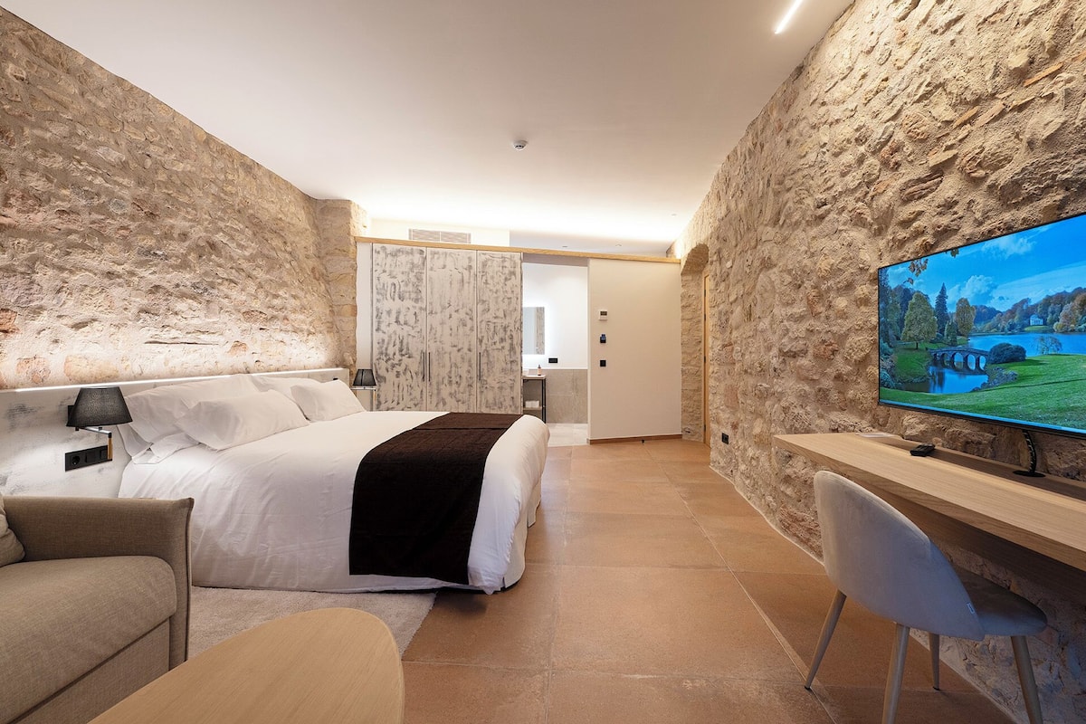 A modern hotel room with a stone wall, a bed, a desk, and a TV.