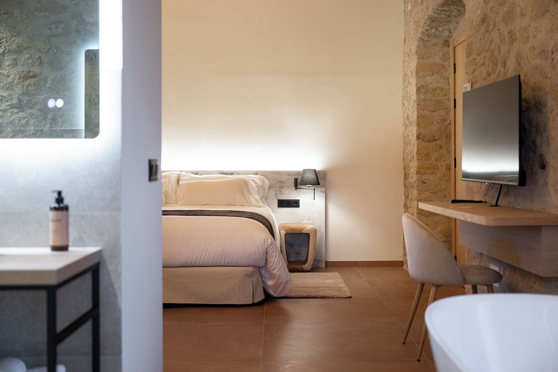 Un dormitorio con cama, mesita de noche, lámpara y televisor montado en una pared de piedra.