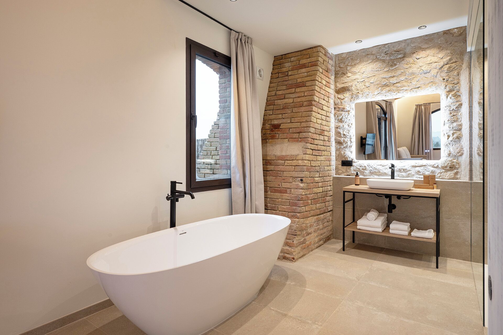 Un baño moderno con una bañera independiente, una pared de acento de piedra, un tocador con espejo y una ventana con cortinas.