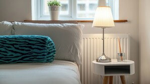 Une chambre à coucher bien rangée, avec un lit, une lampe de chevet, une petite table avec une lampe et une fenêtre donnant sur des immeubles.