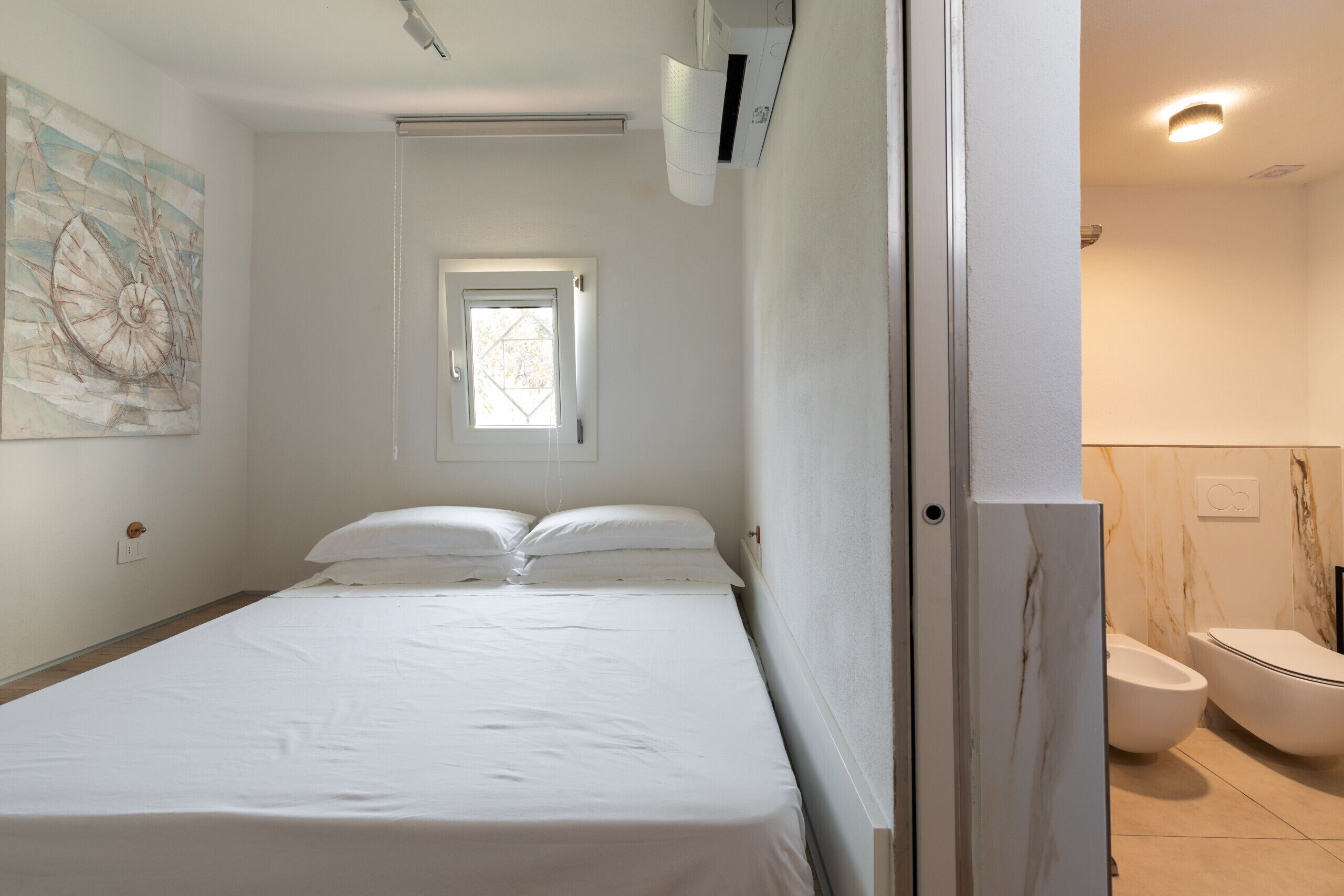 A bedroom with a bed, a window, a door, and a bathroom with a toilet and sink.