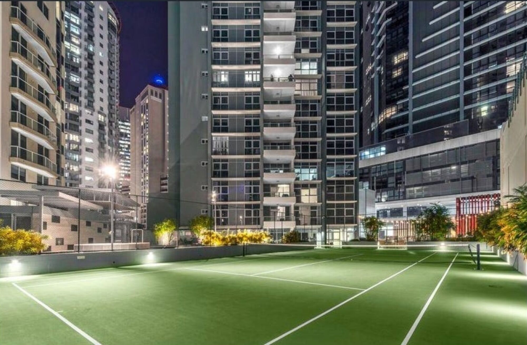 A tennis court surrounded by high-rise buildings at night.