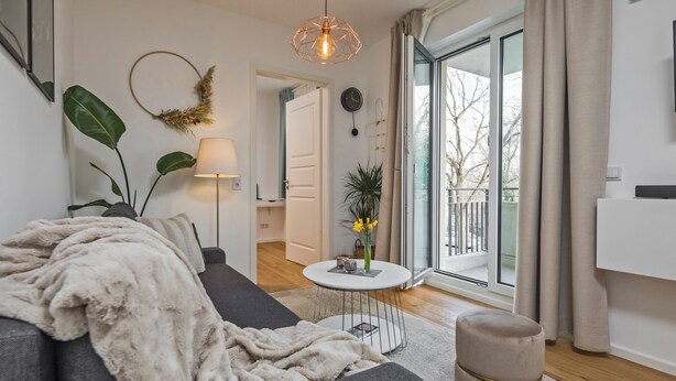 A modern living room with a sofa, a round table, a lamp, and a view of the outdoors. - Gleam Homes Dresden HafenCity Urban District (Dresden)