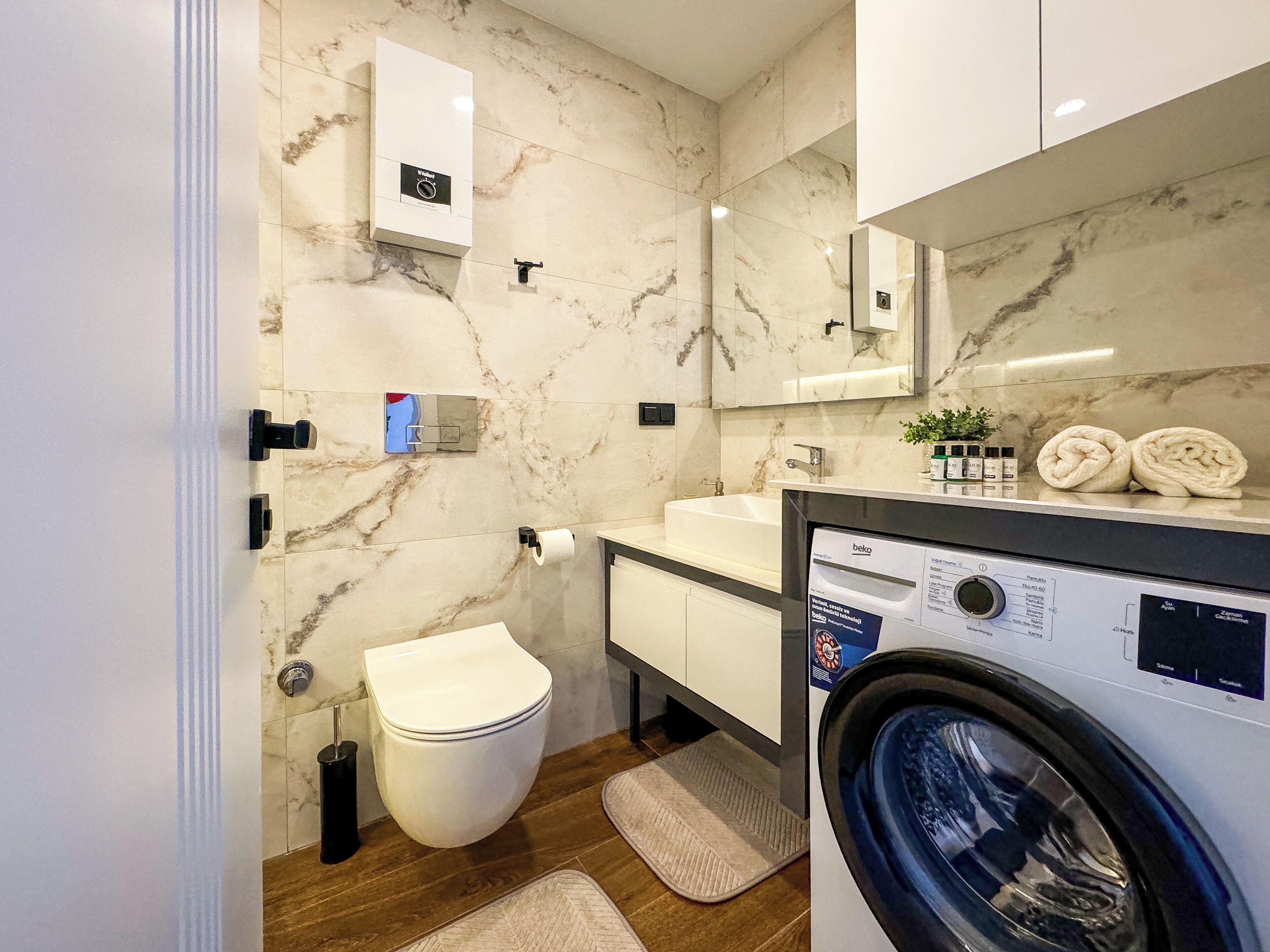 A modern bathroom with a marble wall, a toilet, a sink, a mirror, a washing machine, and a towel rack.