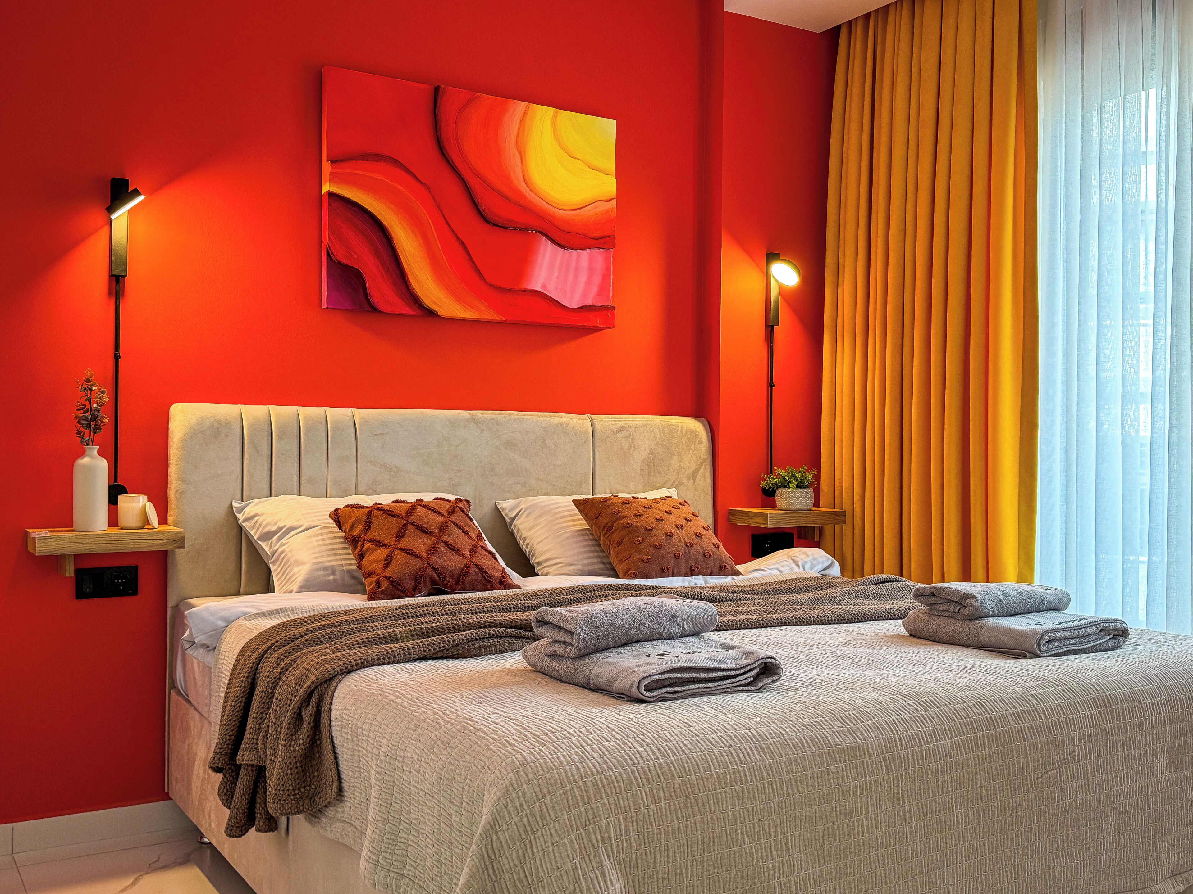 Camera da letto con una parete rossa, una testata beige e un'ampia finestra con tende gialle. Si notano due comodini con lampade, un letto con numerosi cuscini e una coperta.