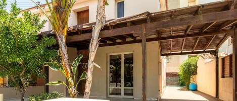 A covered outdoor area with a wooden pergola, a palm tree, and a building entrance with glass doors.