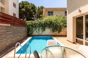 A swimming pool with a glass table and chairs, surrounded by a wooden fence and a building with a tiled exterior.