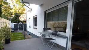 Patio esterno con tavolo e sedie, una porta scorrevole in vetro e un piccolo giardino.
