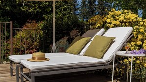 Une terrasse en bois avec deux chaises longues, dont une est ombragée par un parasol beige, le tout entouré d’une végétation luxuriante et de fleurs jaunes.